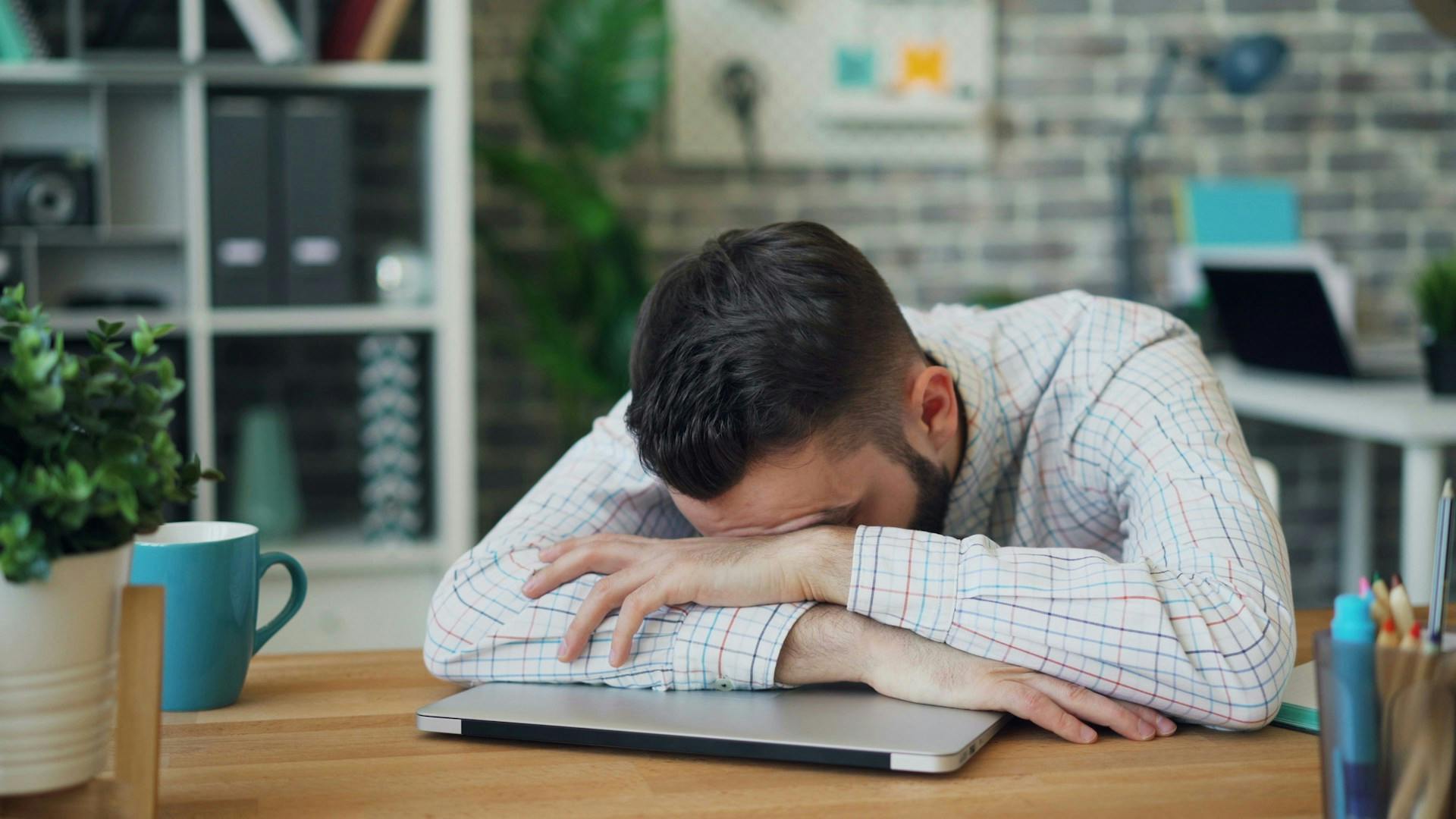 A man slumped over his laptop on a table feeling great stress