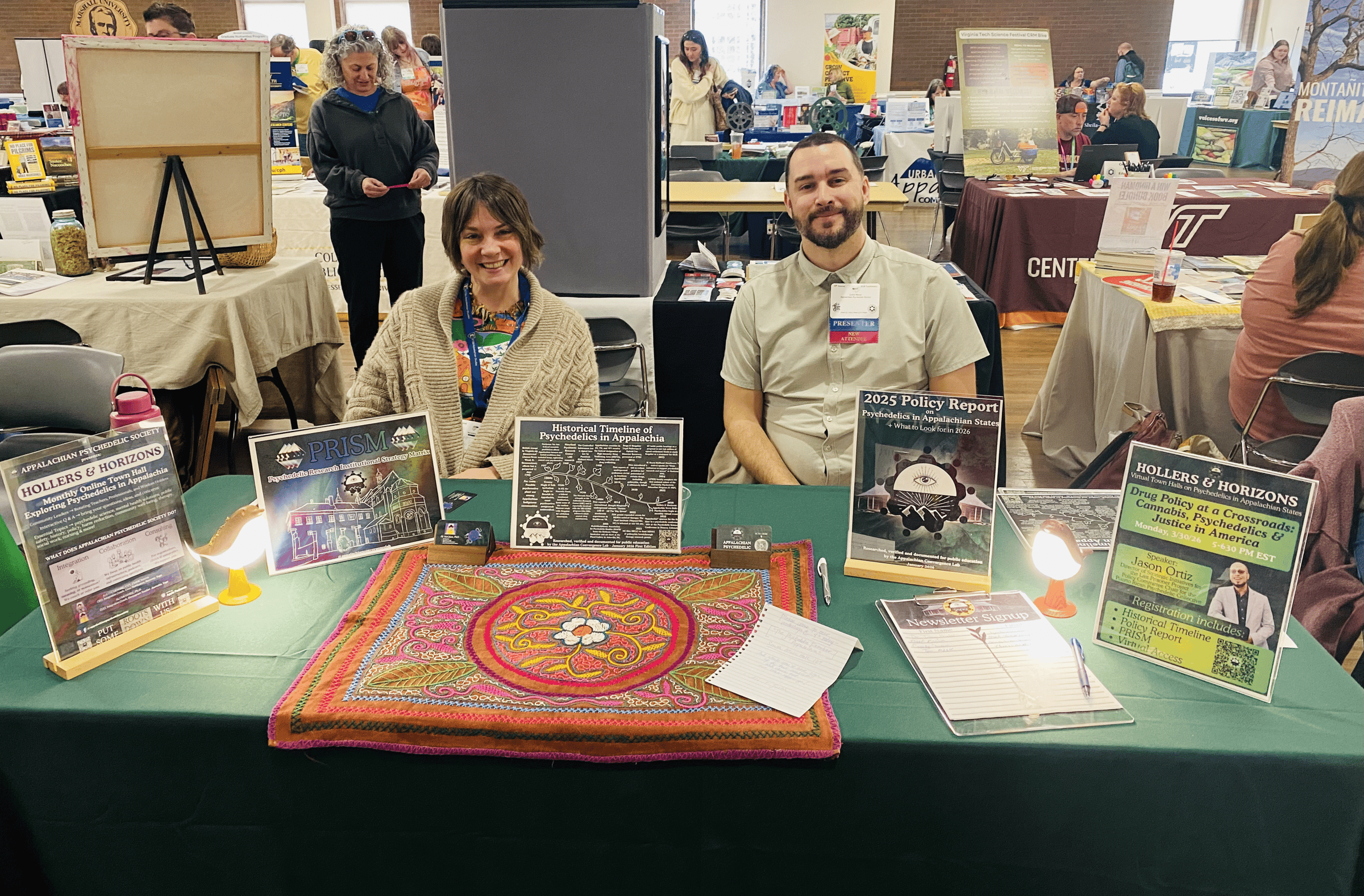 Two people sit smiling behind a table at a conference exhibition hall
