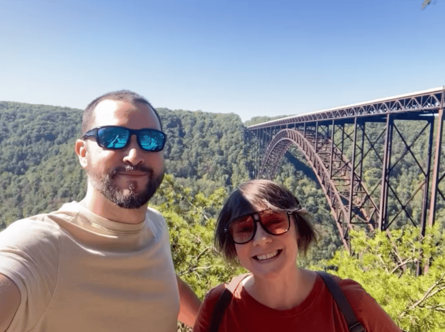A man and woman smile in front of a bridge
