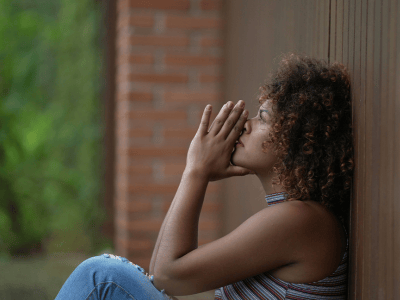 Woman with curly hair sitting against a wooden wall, eyes closed and hands pressed together in front of her face, appearing deep in thought or prayer.