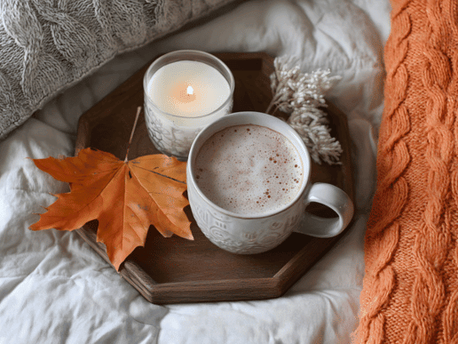 cozy setting with an orange leaf, a candle and a mug of cocoa on a brown wooden try sitting on a bed next to an orange sweater blanket