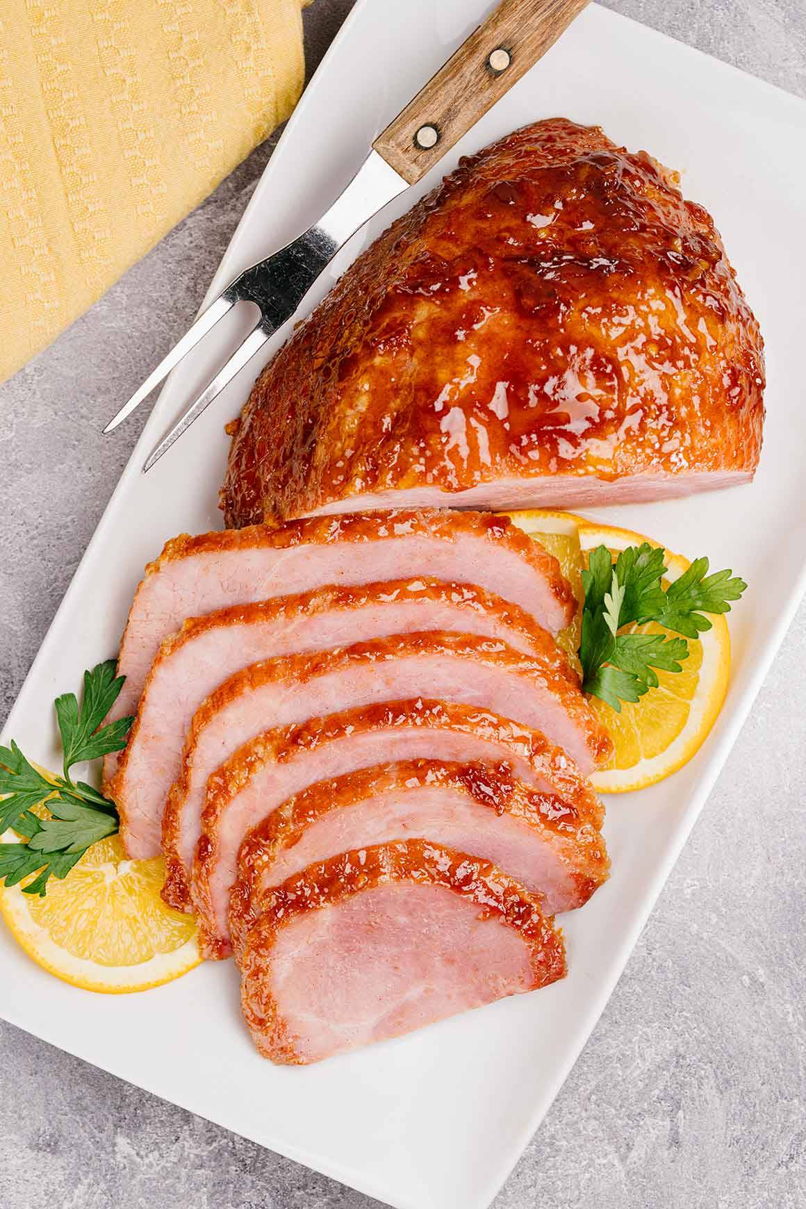 Sliced ham with a glossy orange marmalade glaze on a white platter with decorative orange slices and parsley sprigs.