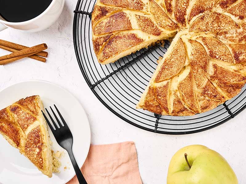 Old-fashioned apple cake with cinnamon-sugar apple slices, a wedge served on a plate, and coffee alongside.