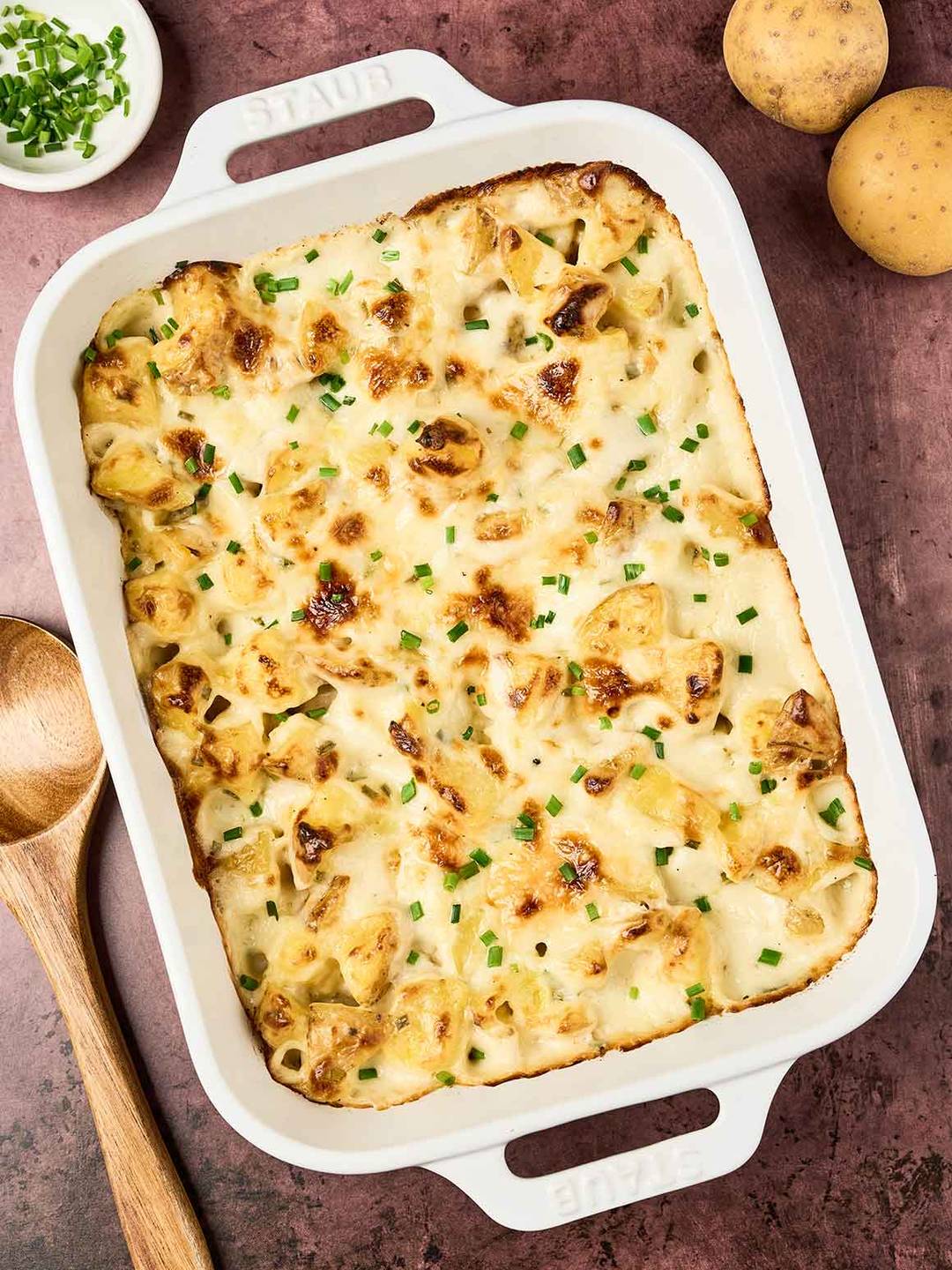 Golden-browned cottage cheese potato casserole in a white baking dish, topped with melted cheese and chopped chives, on a rustic surface with whole potatoes and a wooden spoon nearby.