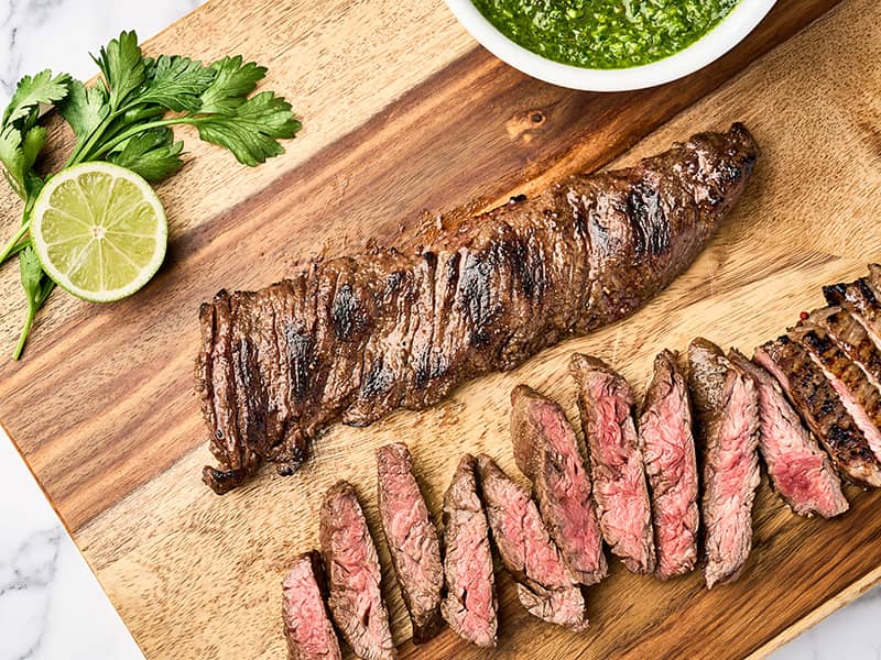 Sliced, grilled skirt steak on a wooden board with a bowl of chimichurri sauce for serving.