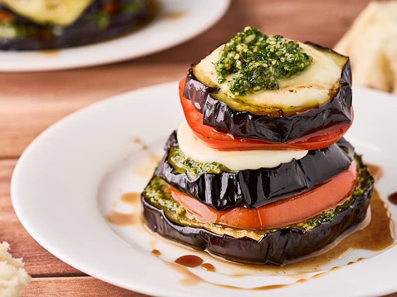 Eggplant Napoleons with pesto, tomato, and fresh mozzarella layered on a white plate and drizzled with balsamic glaze.