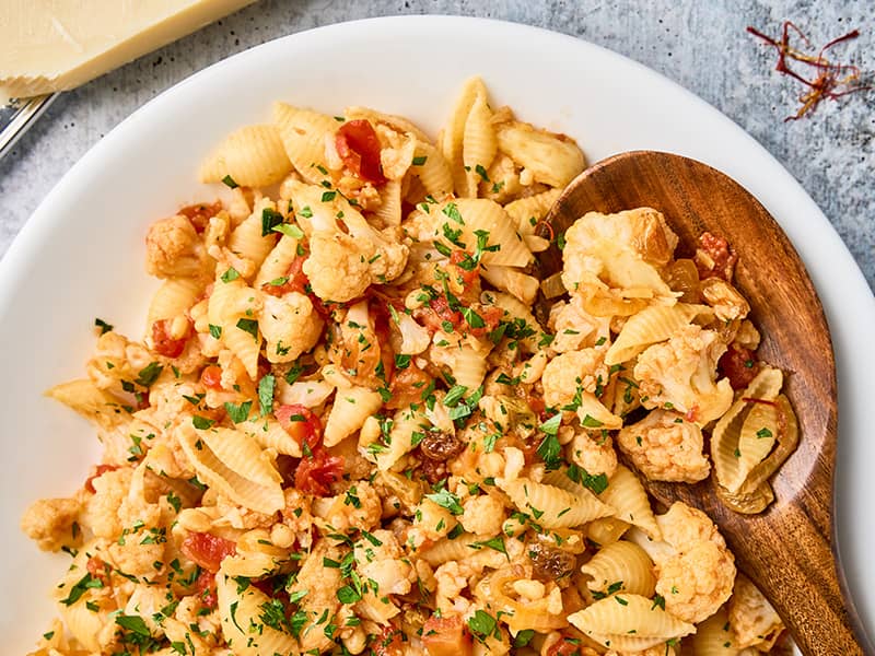 Close-up of Sicilian pasta with cauliflower, shell pasta, tomatoes, raisins, pine nuts, and parsley with a wooden serving spoon.