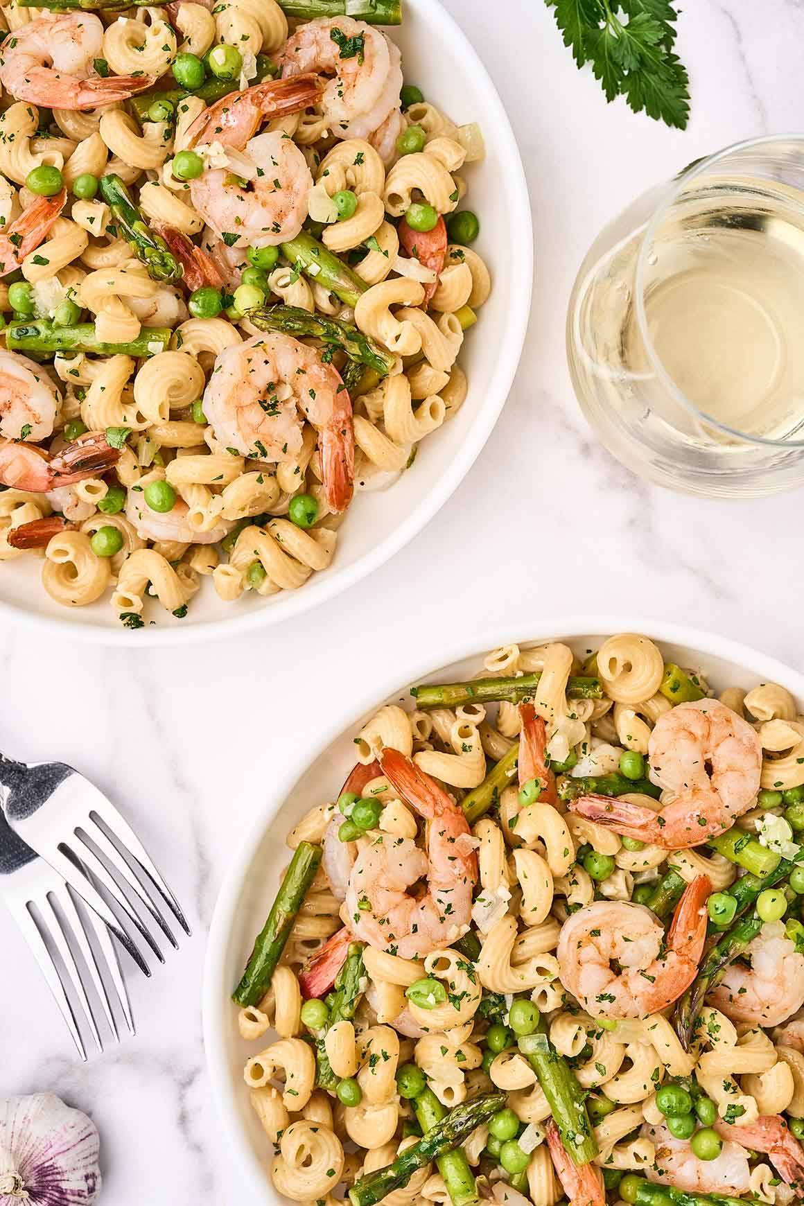 An overhead view of two bowls of shrimp pasta primavera on a marble surface with a glass of white wine, sprigs of parsley and forks.