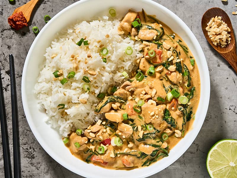 Thai peanut curry with chicken and spinach in a sauté pan beside jasmine rice, scallions, and chopped peanuts.