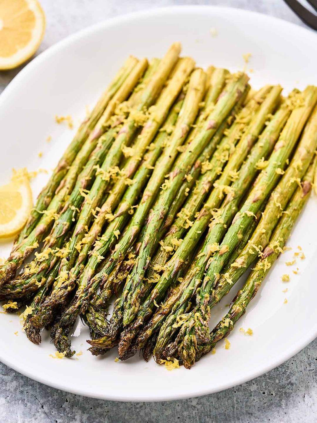 Spears of lemon roasted asparagus topped with lemon zest on a white oval serving plate garnished with lemon slices.