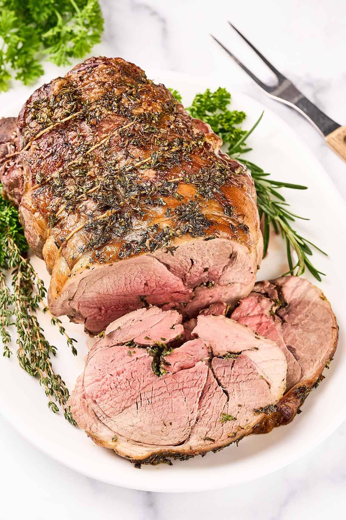 Herb roasted boneless leg of lamb on a serving platter, partially carved and surrounded by fresh herbs.