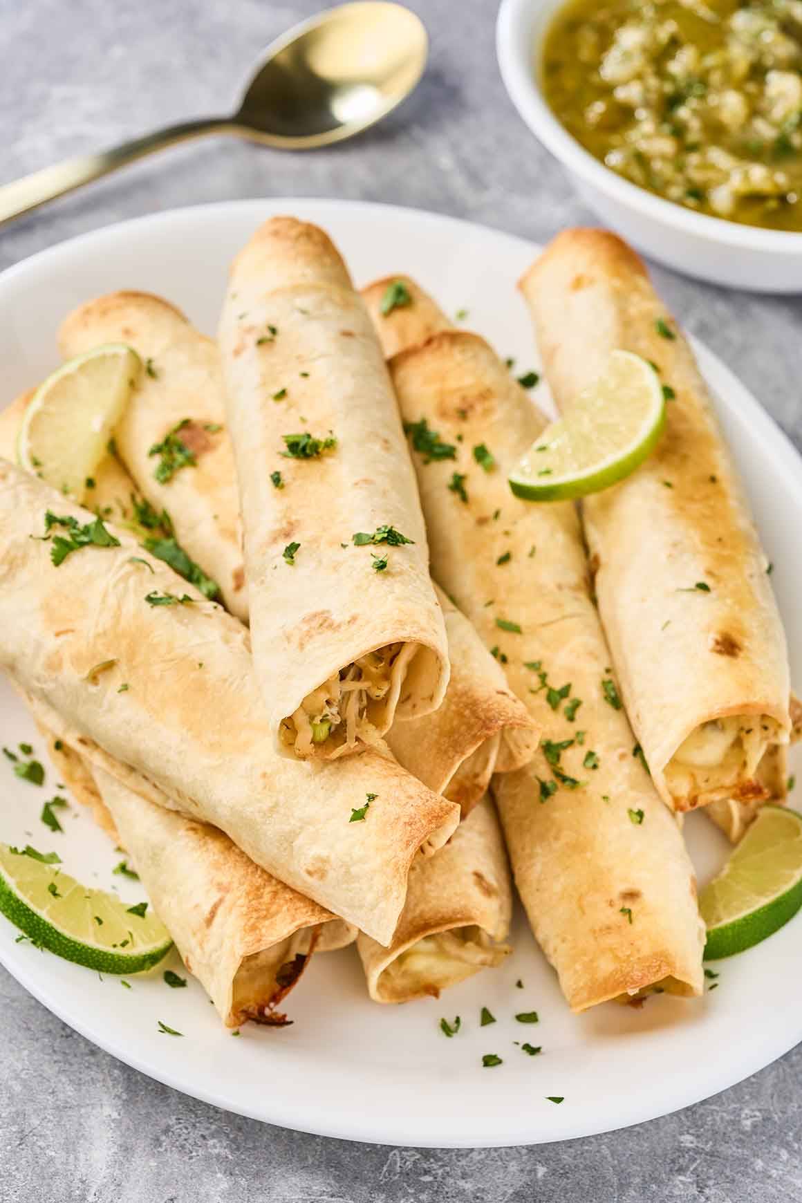 Golden baked chicken flautas on a white platter with lime wedges and fresh tomatillo sauce.