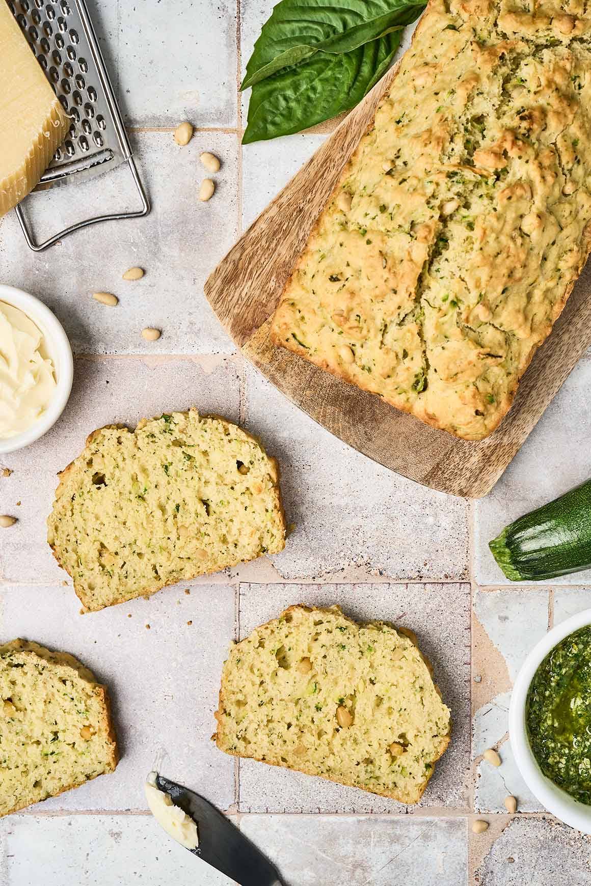 A whole loaf of savory zucchini bread on a wooden board next to fresh slices on the countertop.