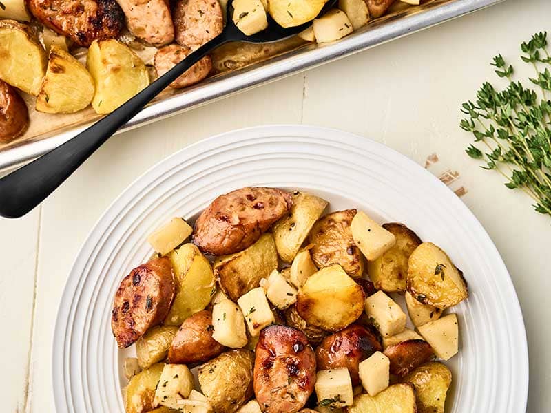 Roasted chicken sausage, potatoes, and apples on a plate, sheet pan in the background.