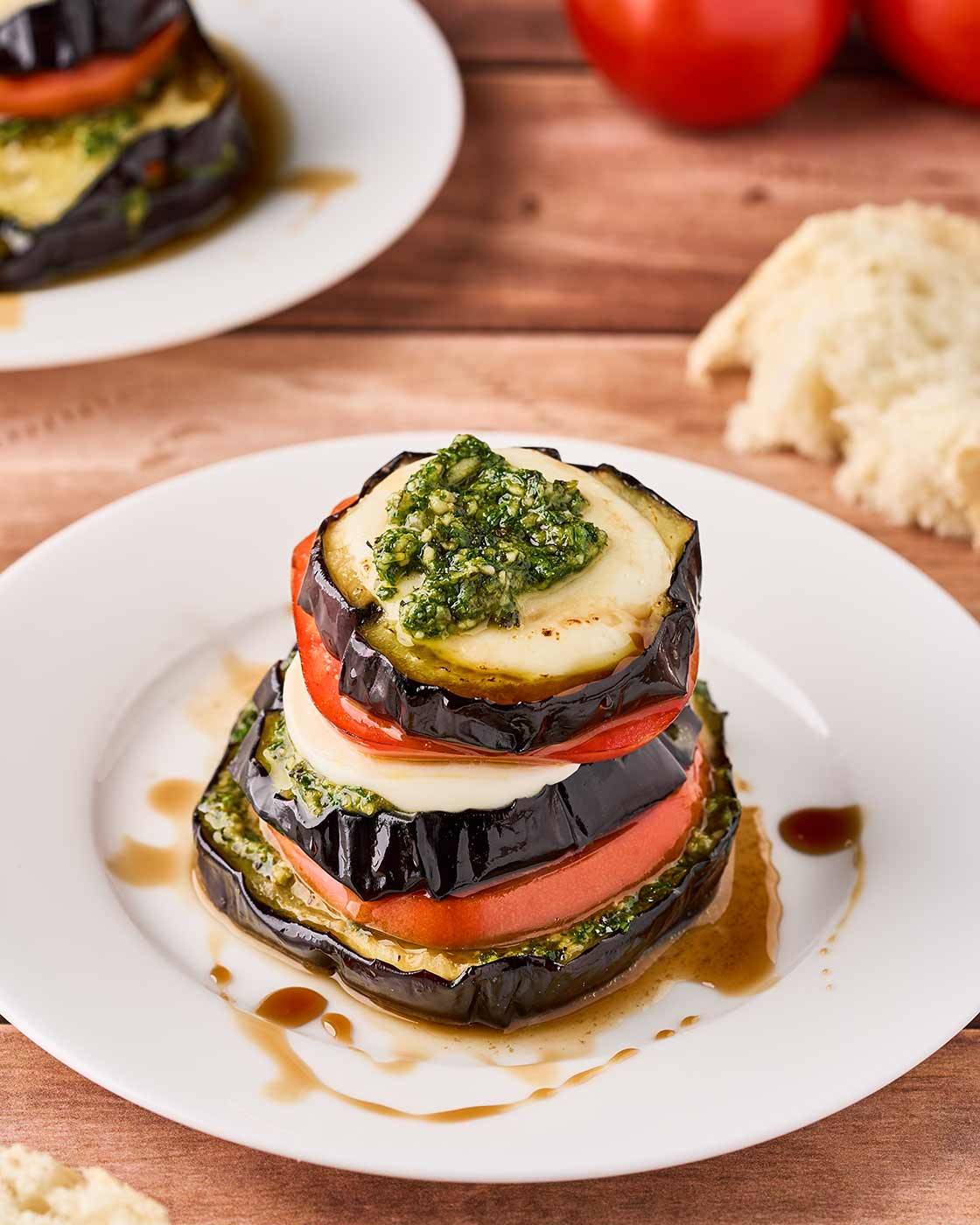 Eggplant Napoleons with pesto, tomato, and melted fresh mozzarella stacked on a white plate with balsamic glaze.