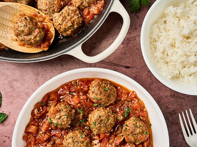 Moroccan meatballs in tomato sauce plated with white rice, with skillet of meatballs and bowl of rice in the background.