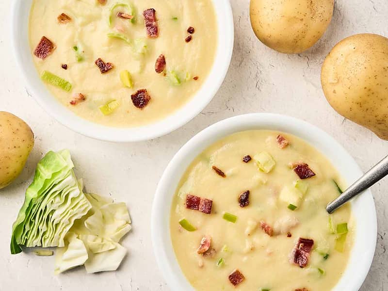 Creamy cabbage leek potato soup topped with bacon and sautéed leeks in white bowls.