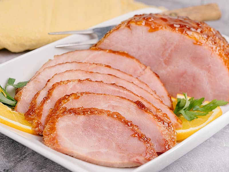 Sliced orange marmalade glazed boneless ham arranged on a white serving platter with orange slices and parsley garnish.