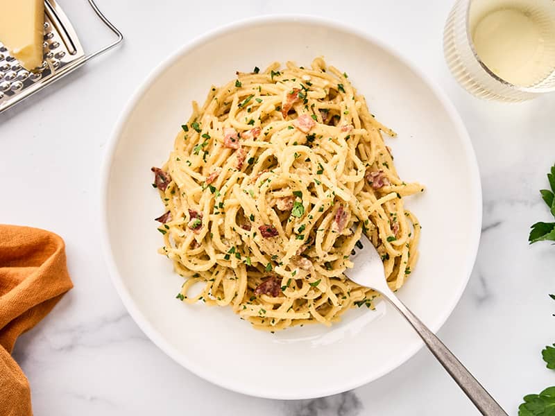 One-pot spaghetti carbonara with bacon, Parmesan, and parsley served on a white plate with fork, wine glass, and fresh parsley.