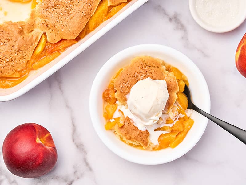 Fresh peach cobbler with biscuit topping and vanilla ice cream, served beside the baking dish.