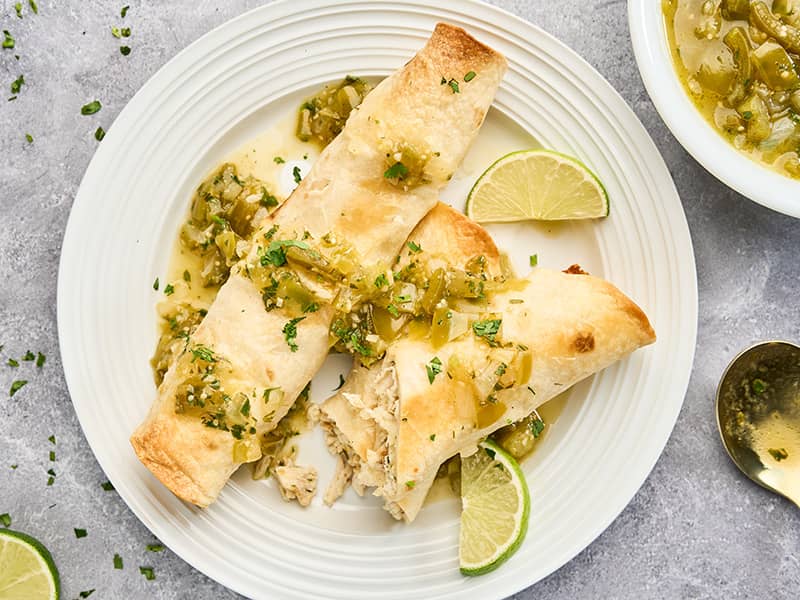 Baked chicken flautas topped with fresh tomatillo sauce and cilantro, with one cut open to show the filling.
