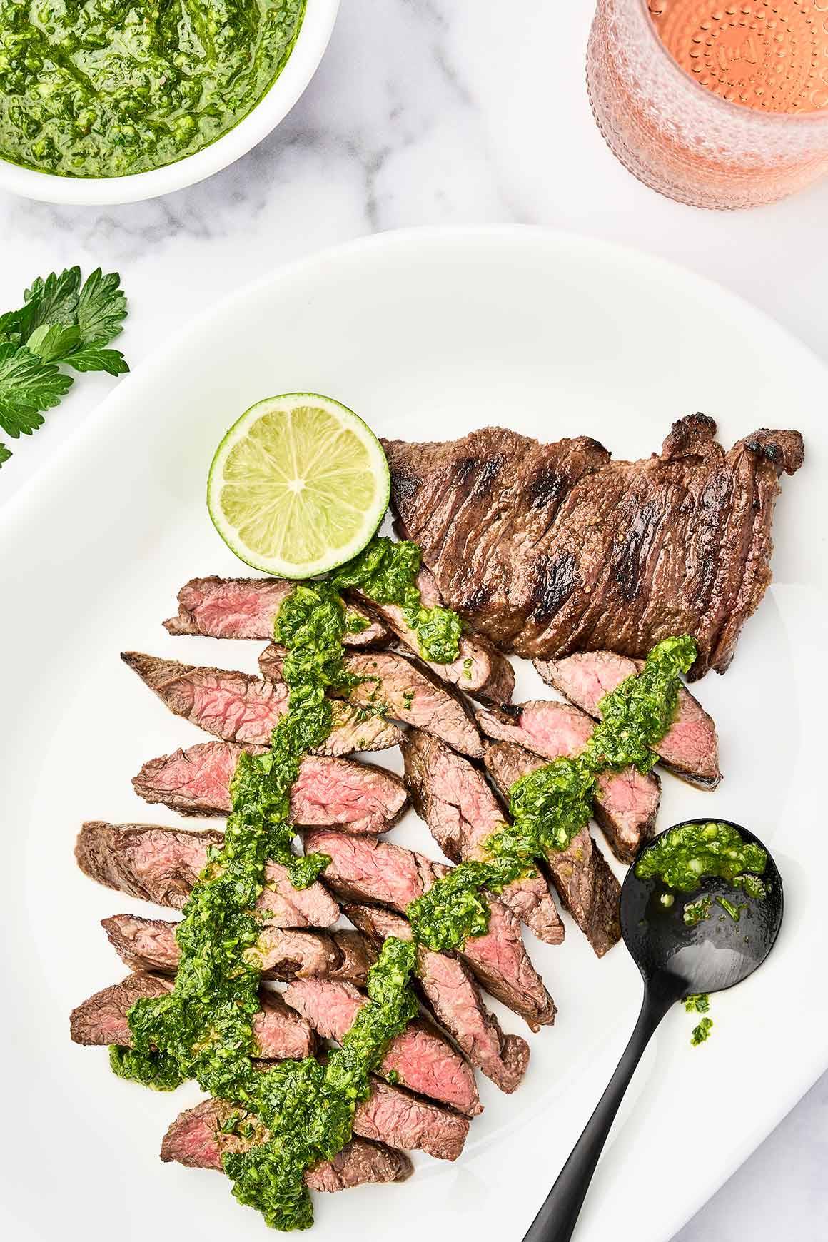 Grilled steak with chimichurri sauce arranged on a serving platter with fresh chimichurri and a lime wedge.