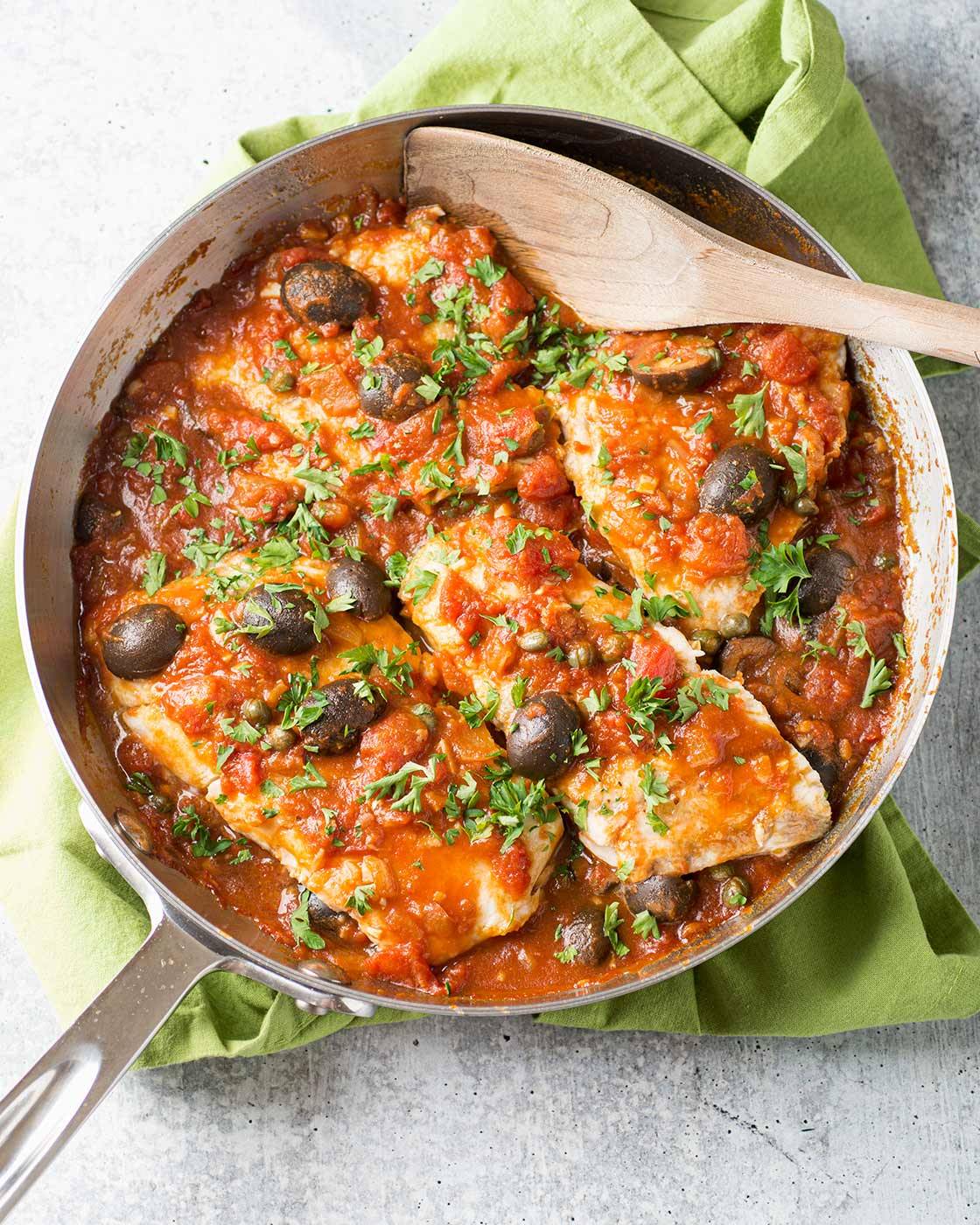 A skillet of white fish fillets in puttanesca-style tomato sauce with kalamata olives, garnished with parsley. 