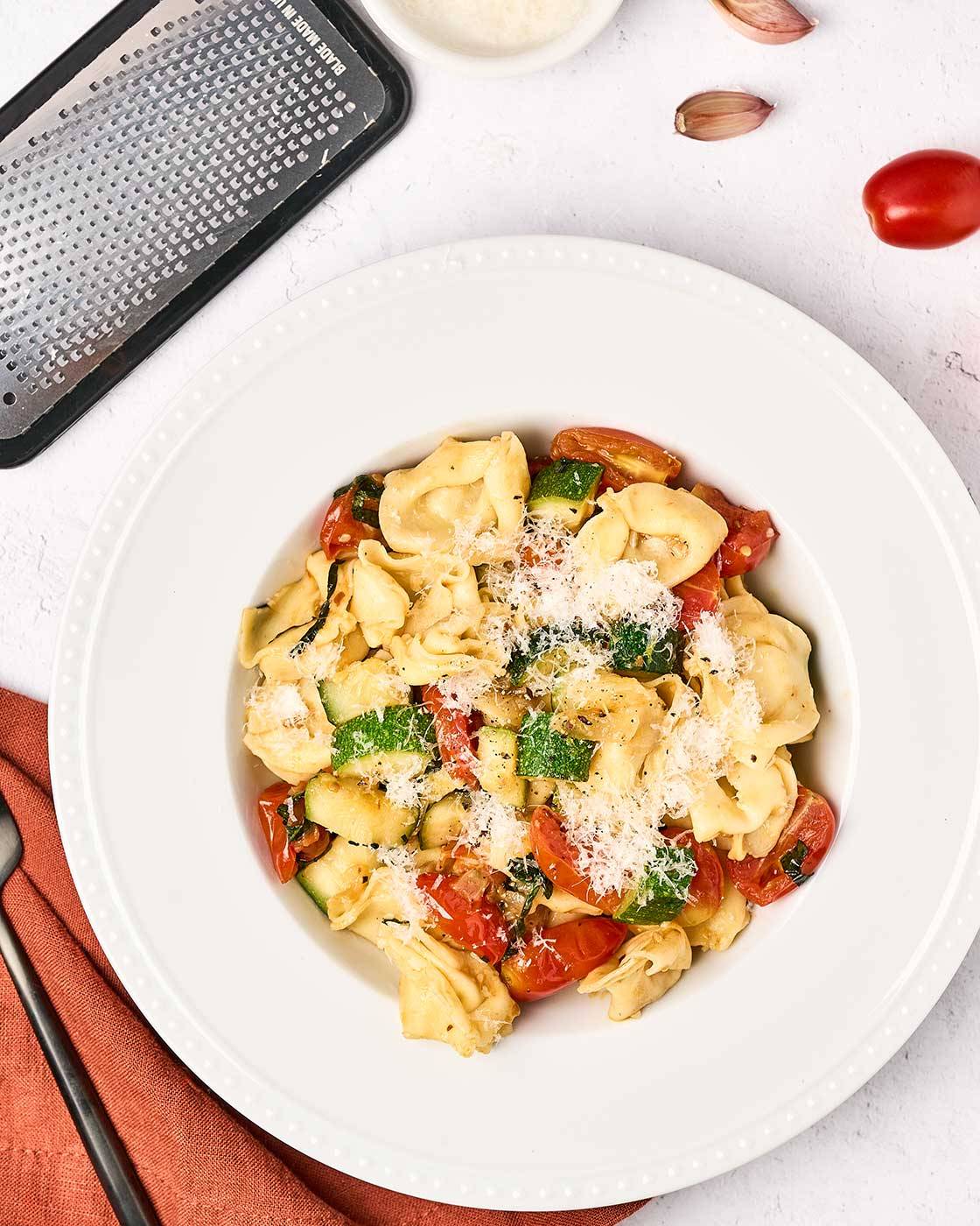 Cheese tortellini with zucchini and tomatoes topped with grated Parmesan, with garlic and cheese in the background.