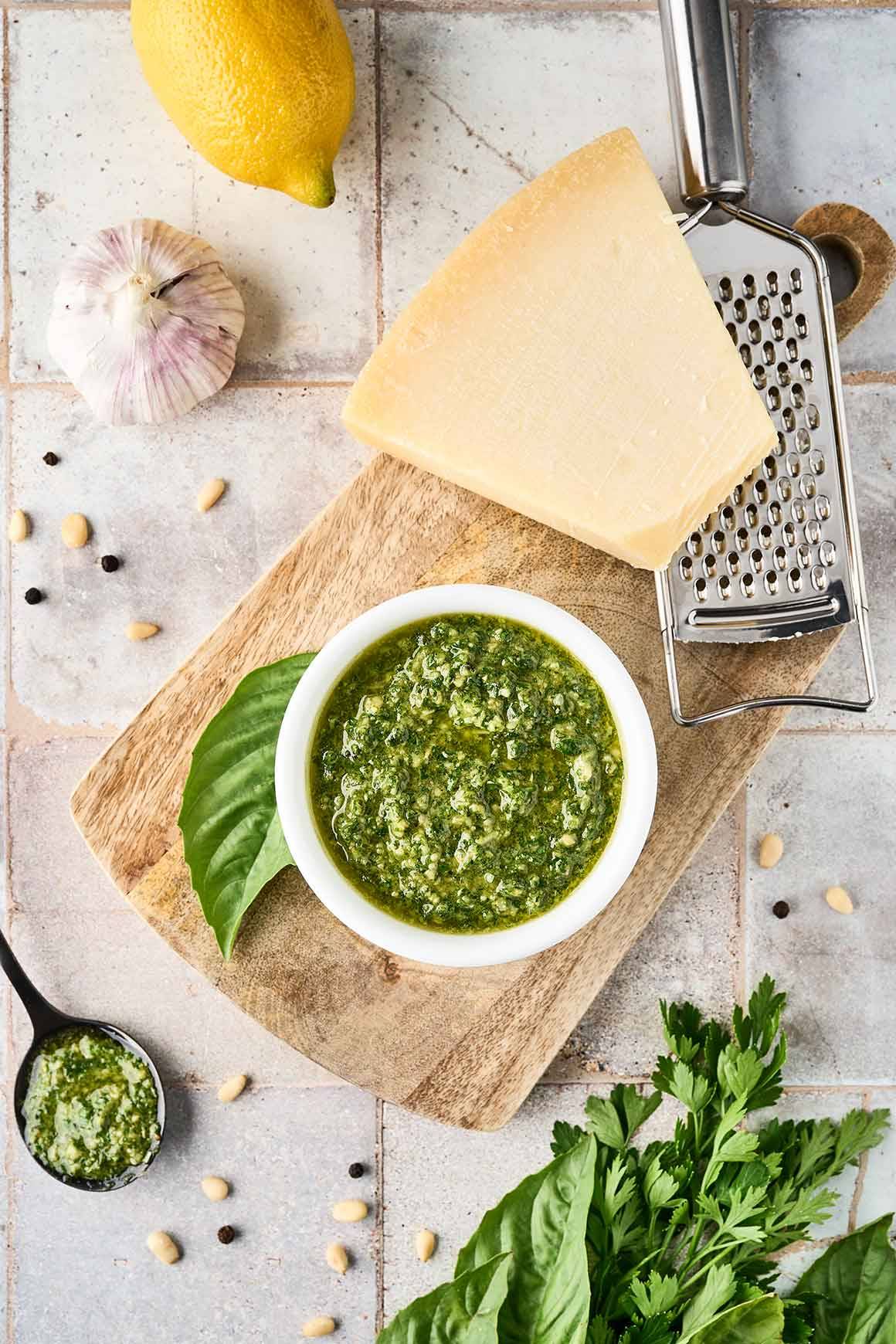 Basil and parsley pesto in a bowl on a wooden board, surrounded by fresh basil, garlic, and a wedge of Parmesan.