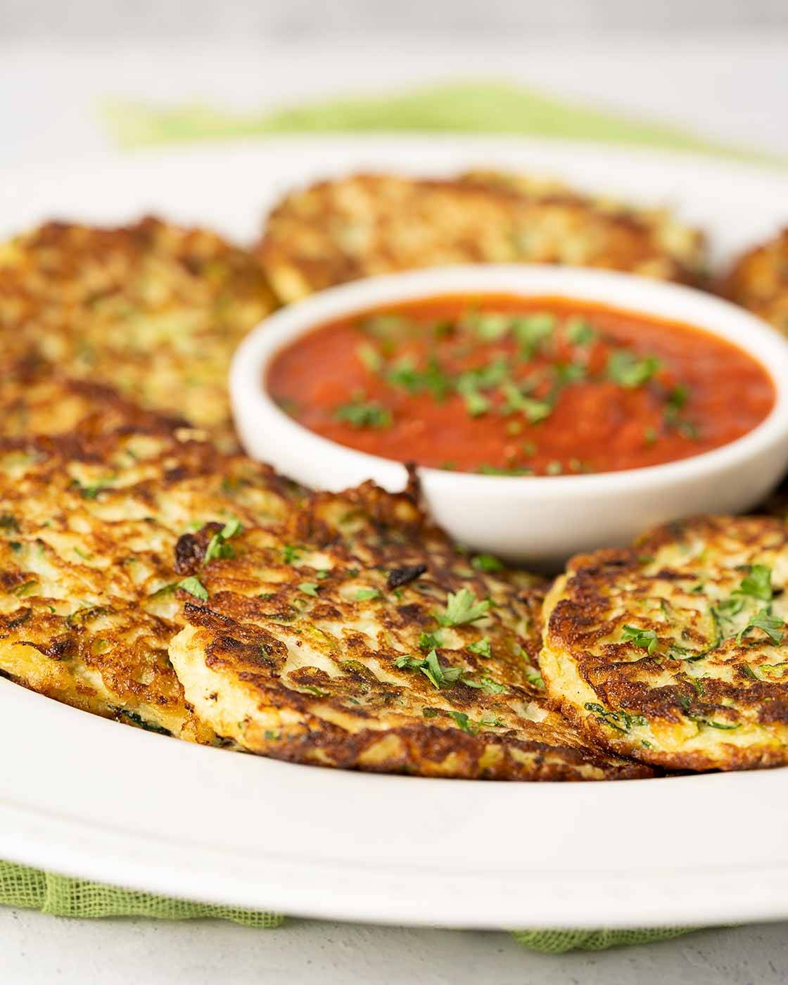 A serving platter of golden zucchini fritters and a bowl of marinara sauce for dipping.