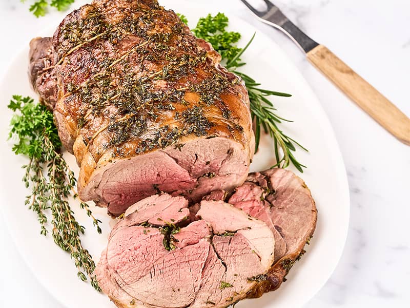 Herb roasted boneless leg of lamb on a serving platter, partially carved and garnished with fresh herbs.