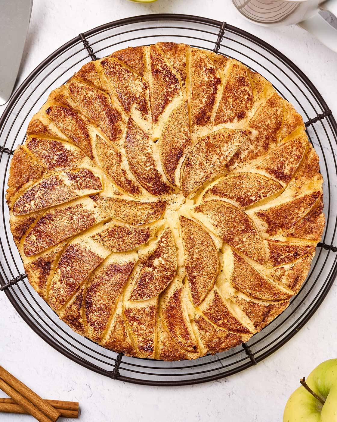 Old-fashioned apple cake topped with cinnamon-sugar apple slices on a cooling rack, with a slice served on a plate.