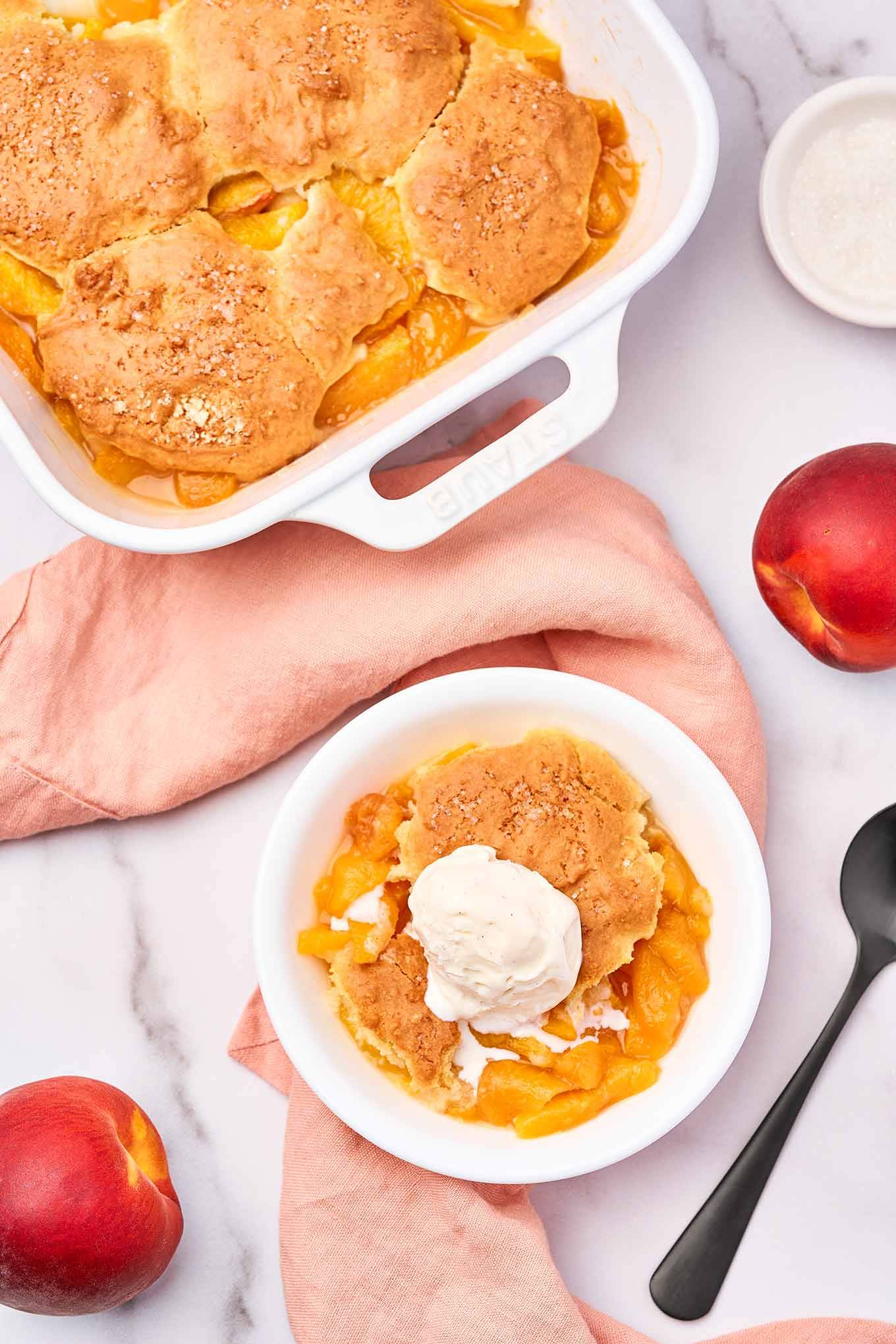 Fresh peach cobbler with biscuit topping and vanilla ice cream served in a white bowl, viewed from above.