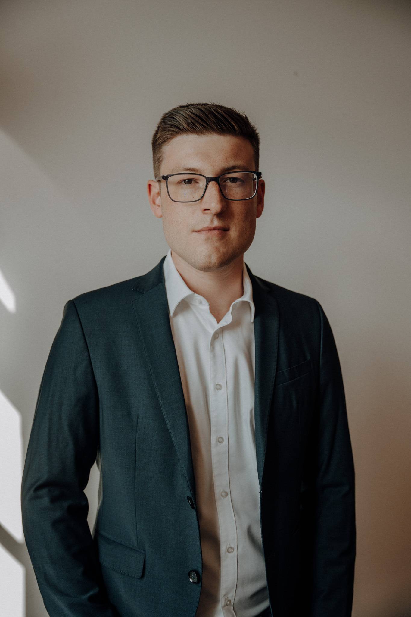 A man with short hair and glasses is wearing a white shirt and a dark blazer. He is standing against a plain background, looking directly at the camera with a neutral expression.
