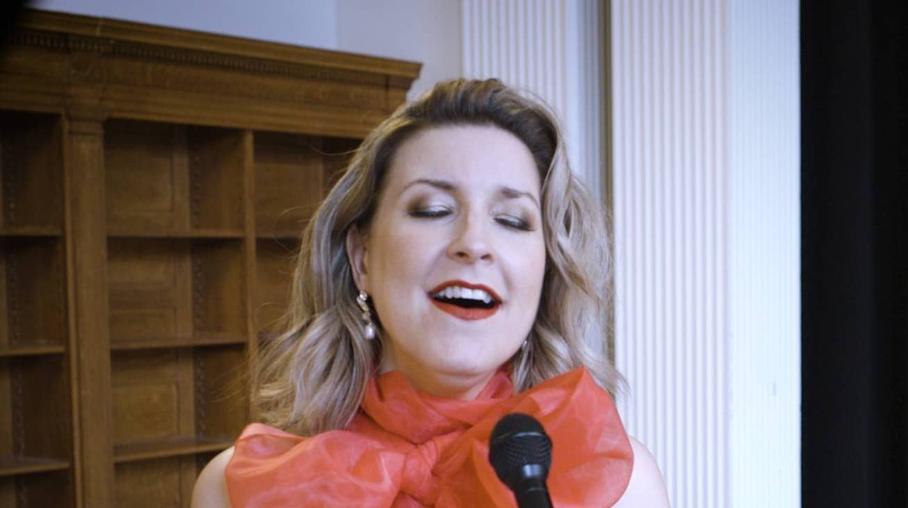 A woman is singing into a microphone, wearing a sheer red top with a large bow at the neck. Her eyes are closed and she appears to be passionately performing. The background includes wooden shelves and white columns.