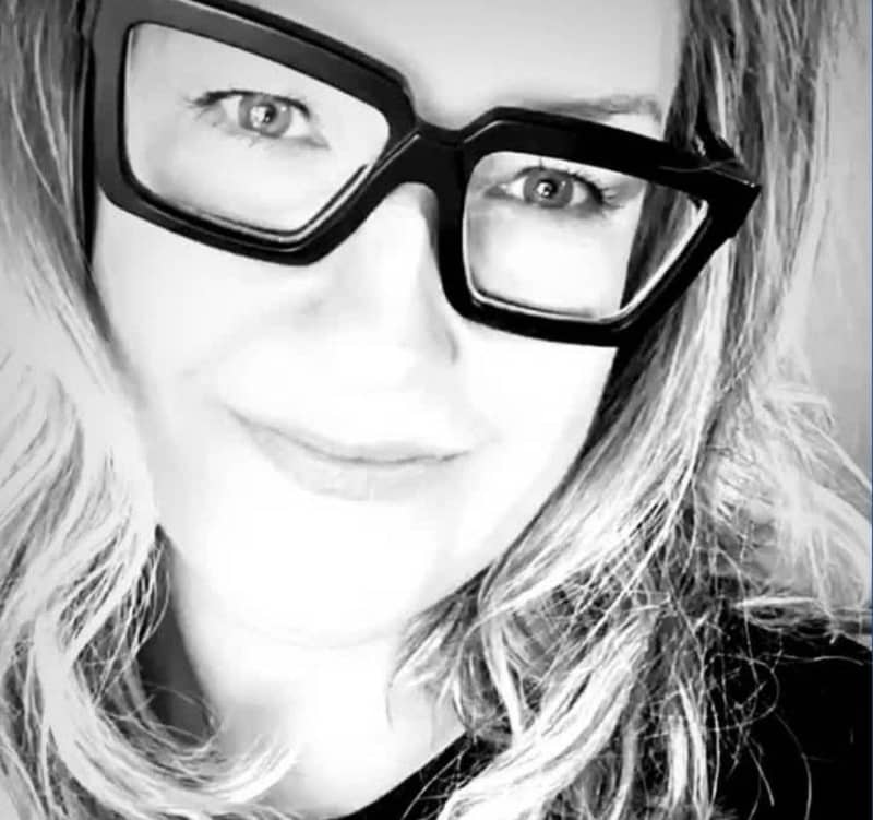 A black and white close-up image of a person with long, wavy hair wearing large, thick-framed glasses. The background is blurred, focusing mainly on the person's face and eyewear.