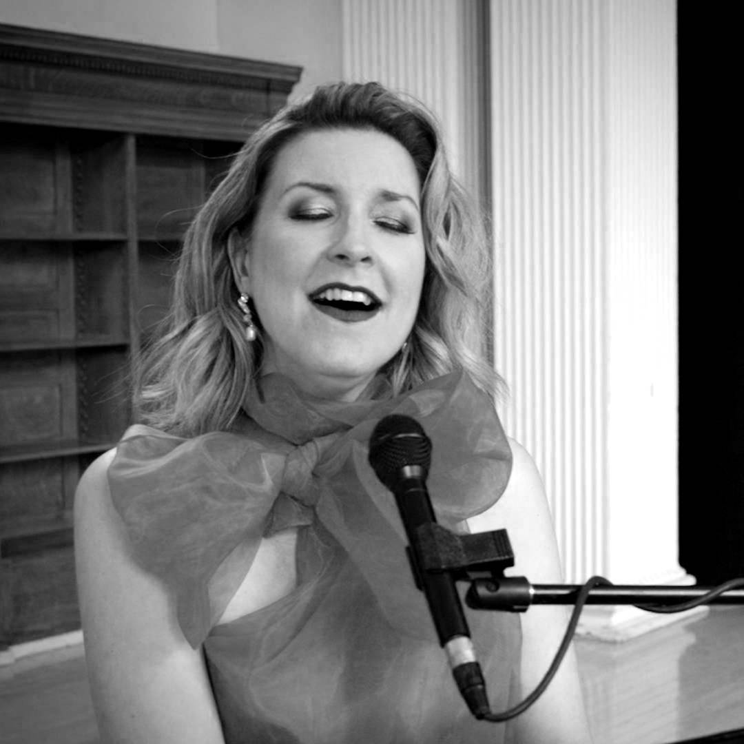 A woman singing into a microphone with her eyes closed. She is wearing a bright red sheer top with a large bow. The background features wooden shelves and white pillars. Her expression suggests she is passionately engaged in the music.