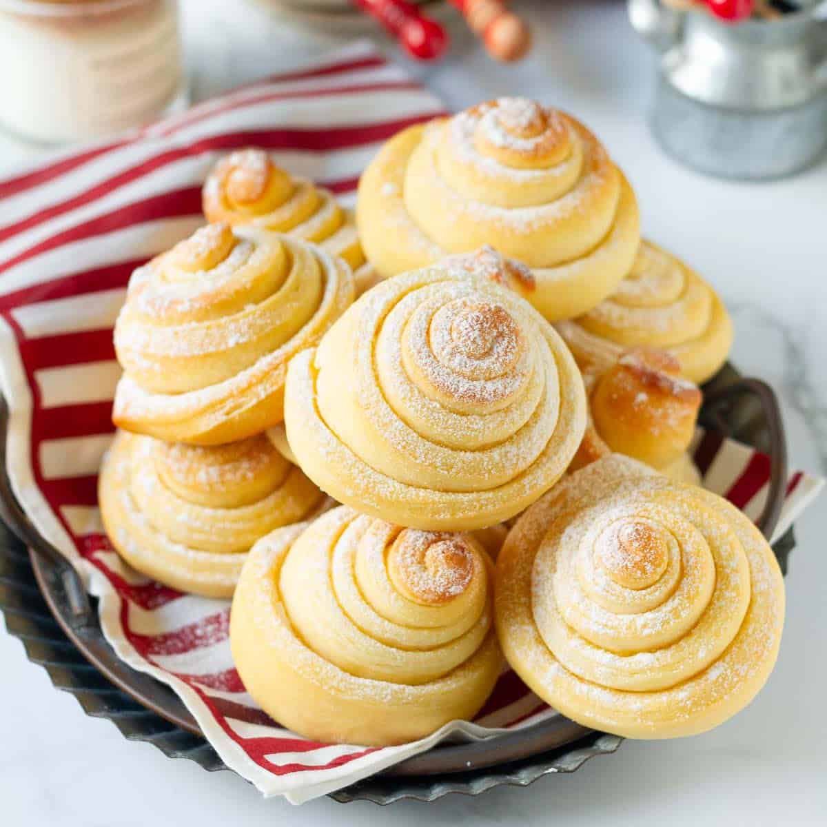 Bread rolls dusted with powdered sugar on a red and white napkin lined tray.
