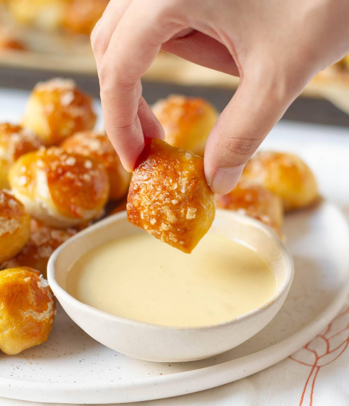 A hand holding a pretzel bite over a bowl of dip.