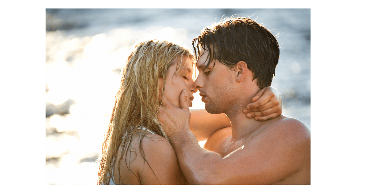 A blonde woman and a dark haired man kissing on a beach.