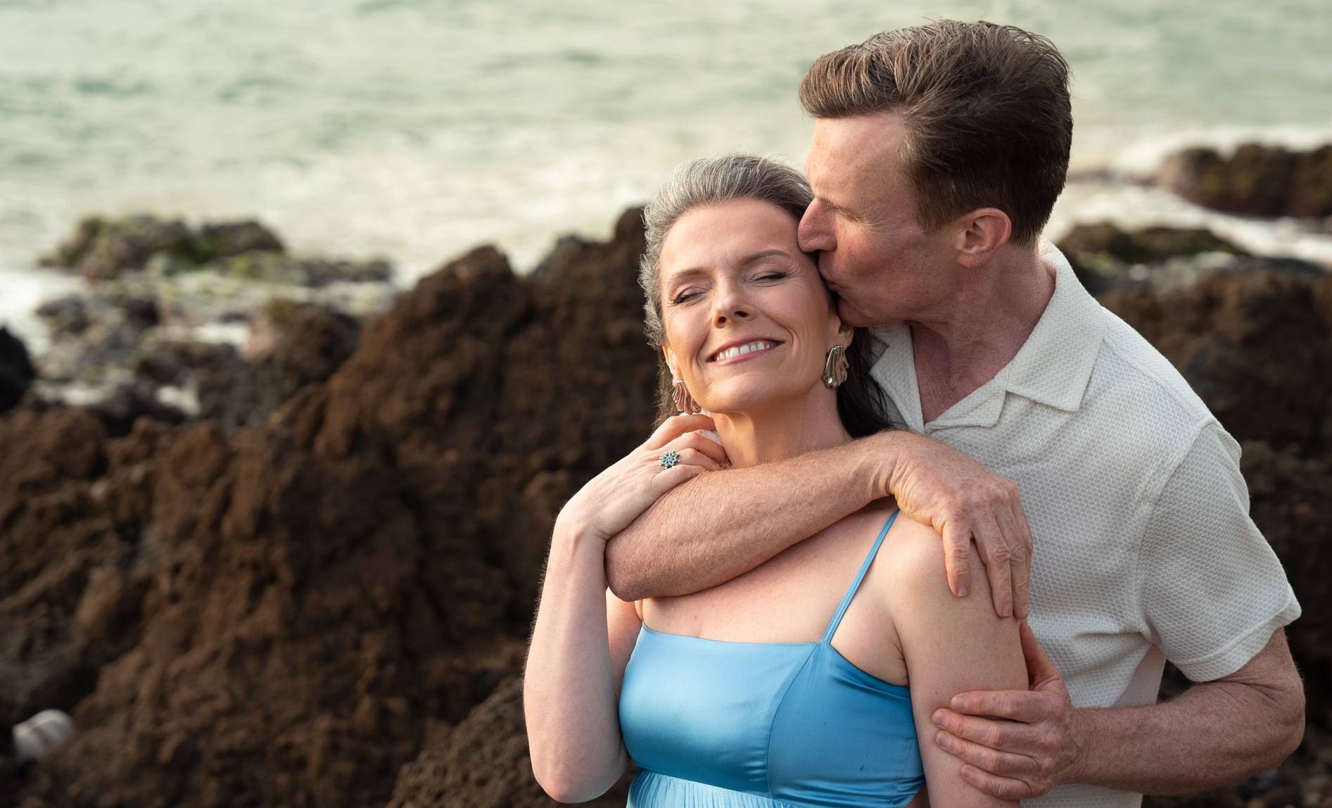 Kurt kisses Michele's temple on a rocky beach