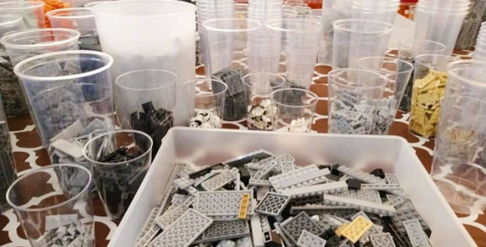 Clear plastic drawer full of various grey LEGO pieces (with some pale yellow ones), surrounded by clear plastic cups of various sizes, with various parts ranging from light grey to black and pale yellow, plus a bucket 3/4 full with LEGO pieces