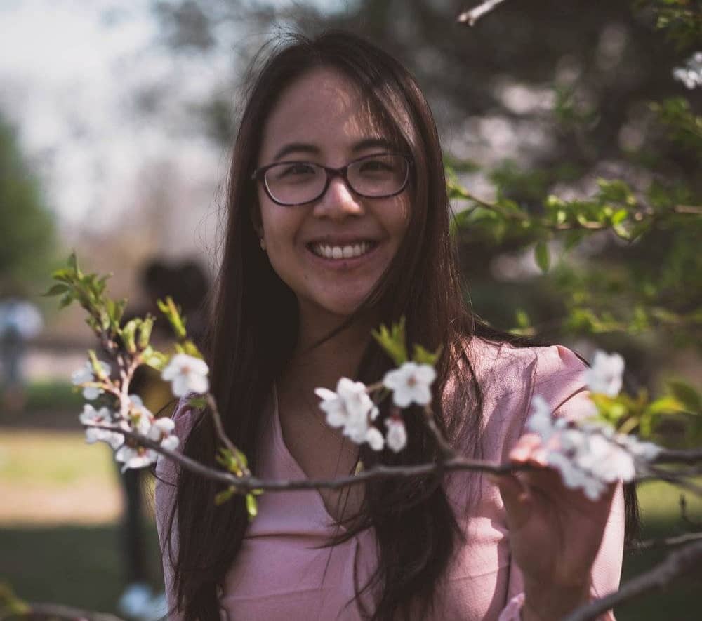 A smiling woman wearing glasses and a light pink blouse stands outdoors, holding a flowering tree branch with white blossoms.