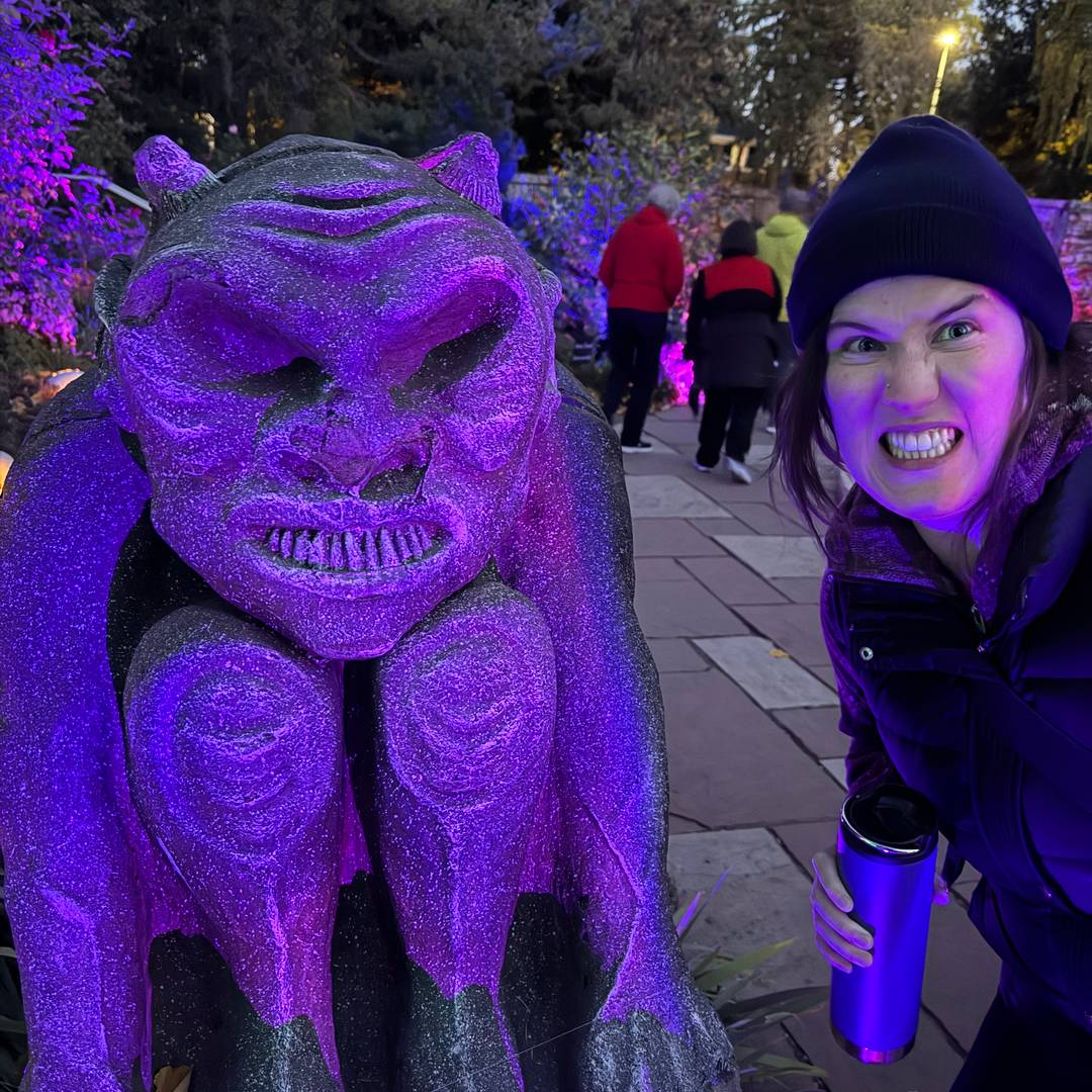 A woman playfully baring her teeth beside a purple-lit gargoyle statue at night, surrounded by people walking through an outdoor Halloween event.