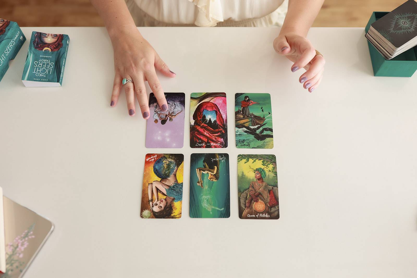 A person is doing a tarot card reading on a white table. Six colorful tarot cards are laid out in two rows of three. The person’s left hand is gesturing toward the cards, and their right hand is resting nearby. Tarot card decks and boxes are visible on the edges of the table.