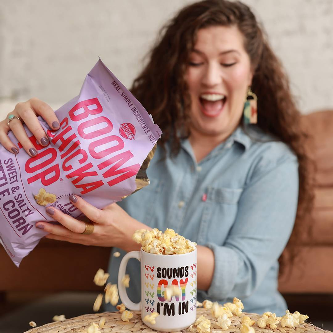 A smiling woman with long curly hair pours popcorn from a bag into a colorful lettered mug, with kernels spilling onto the table around it.