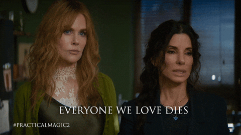 Two women stand side by side in a dim, moody room, looking ahead with serious, wary expressions. The woman on the left has long wavy strawberry-blonde hair and wears a cream lace top. The woman on the right has long dark hair and a dark top. Caption reads “Everyone we love dies,” with the hashtag #PracticalMagic2.