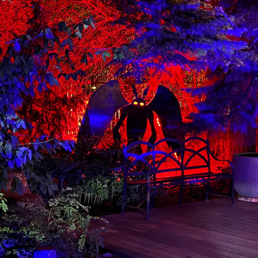 A spooky red and blue light display highlighting a large winged gargoyle statue seated behind a bench amid lush greenery.