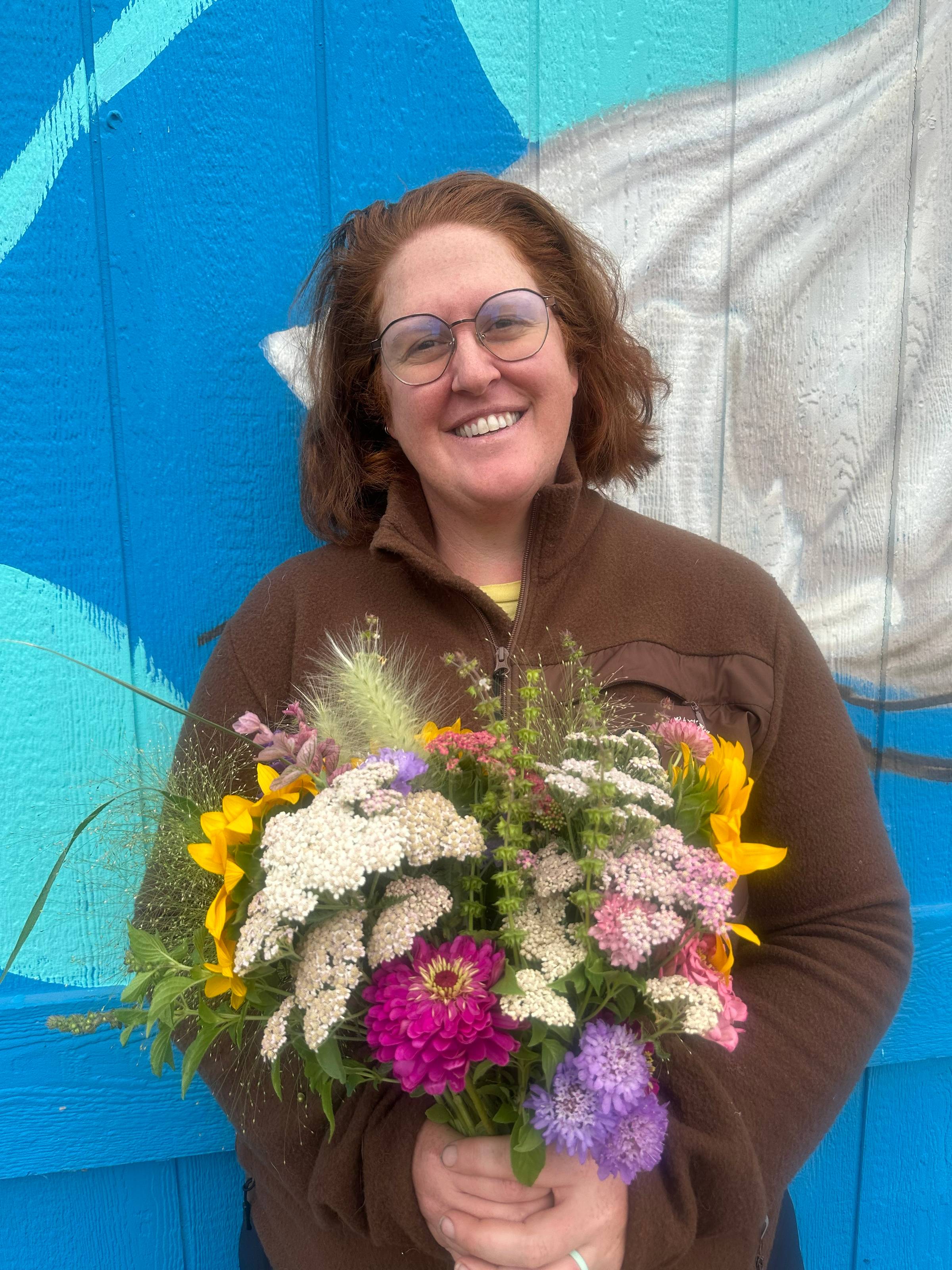 A smiling woman holds a large bucket overflowing with colorful pink, purple, white, red, and orange flowers. She stands in front of a bright blue wall painted with a large floral mural.