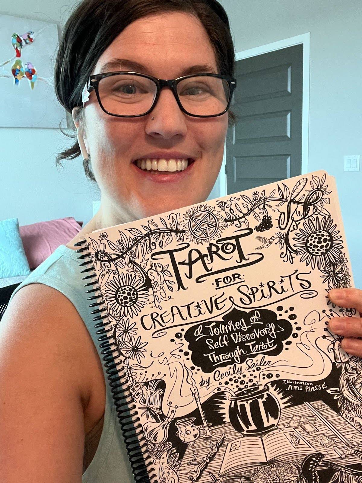 A smiling woman wearing glasses holds up a spiral-bound workbook titled Tarot for Creative Spirits: A Journey of Self-Discovery Through Tarot by Cecily Sailer. The black-and-white illustrated cover features mystical doodles of candles, plants, and a cauldron.
